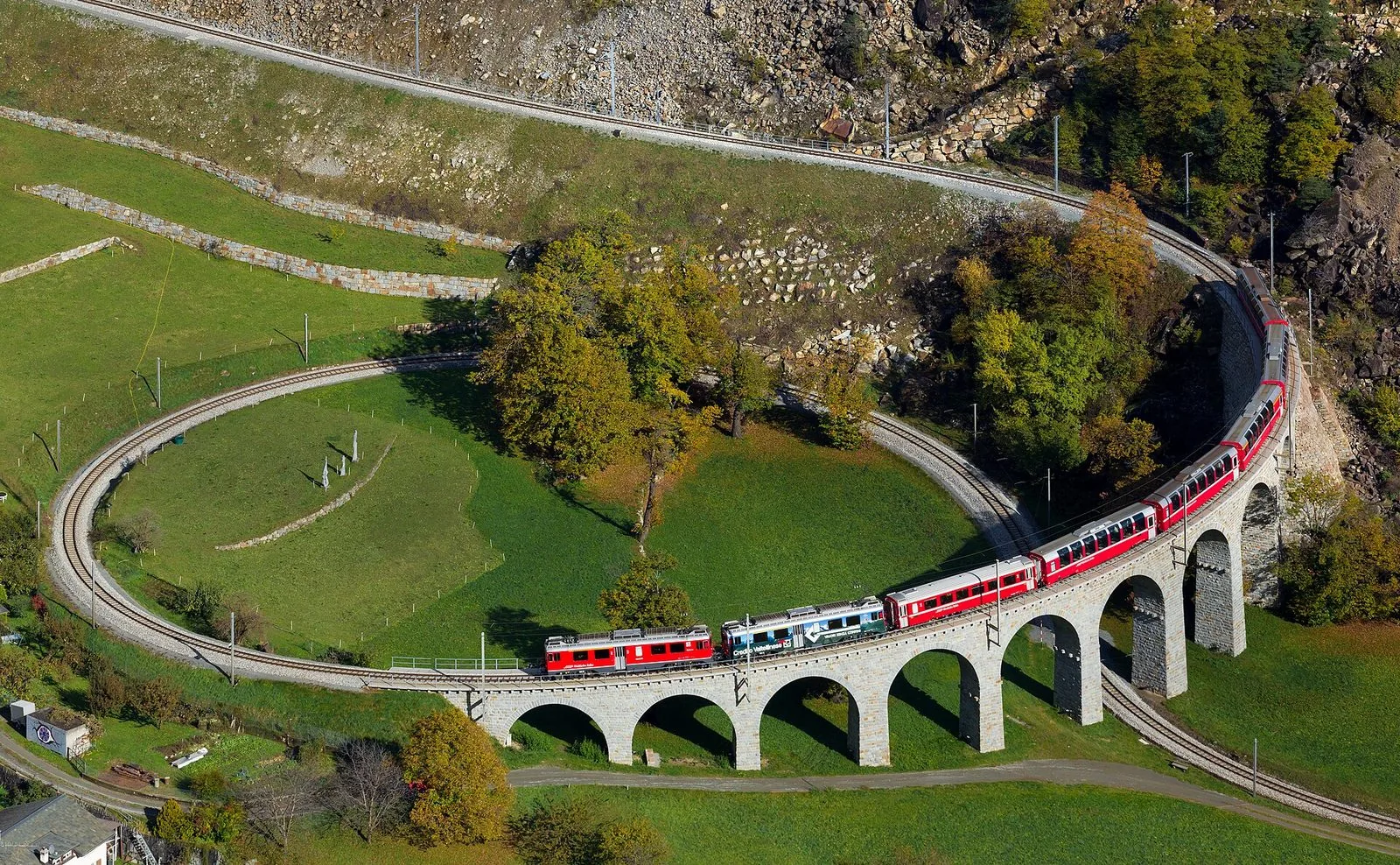 Il Trenino Rosso del Bernina percorre il viadotto elicoidale di Brusio tra prati e alberi autunnali