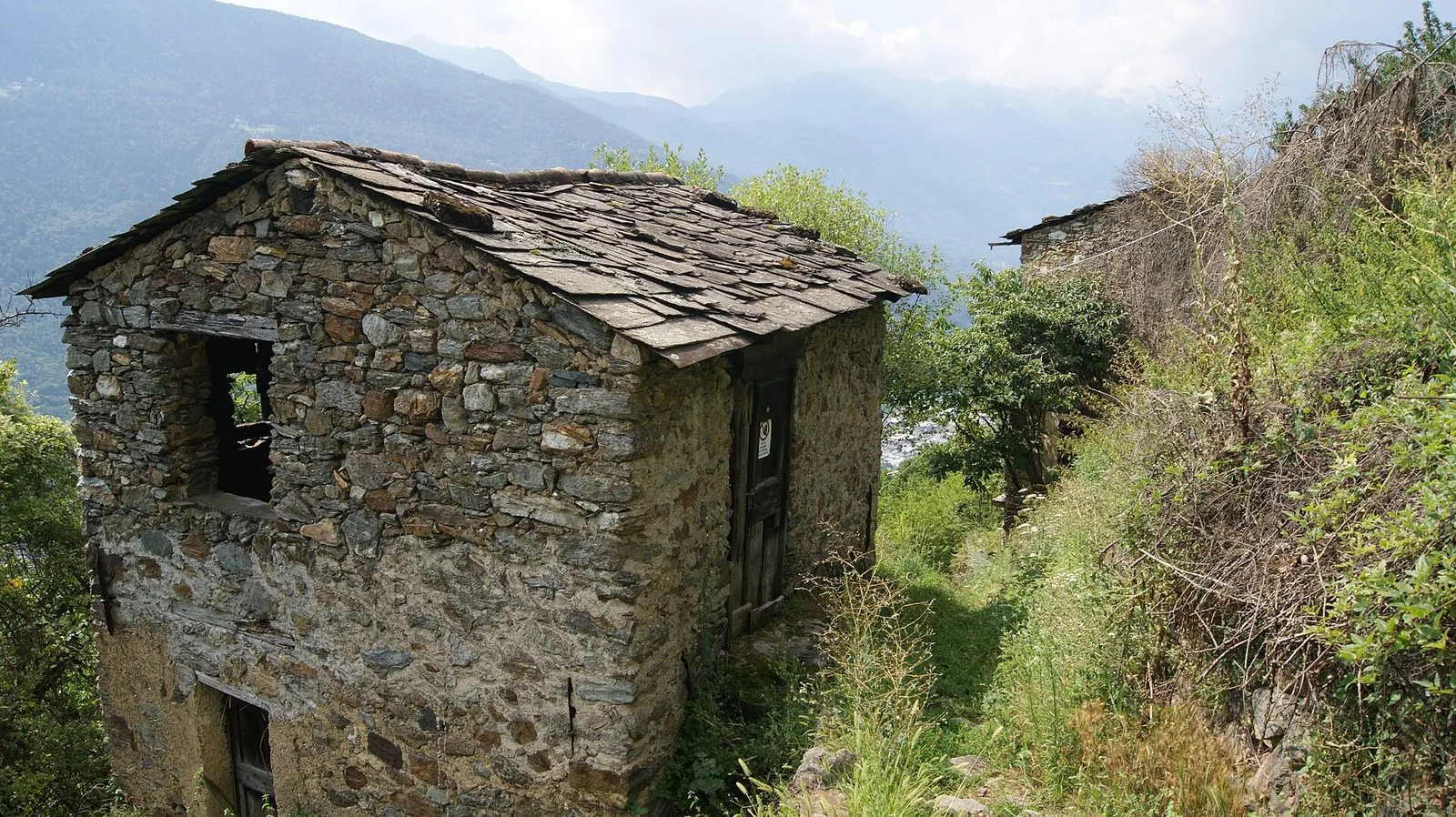 Sentiero escursionistico accanto a una baita in pietra sopra Tirano, con la valle e le montagne sullo sfondo